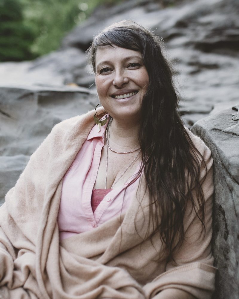 Portrait of Beth Ebers, registered midwife, herbalist and somatic therapist, seated on river rocks